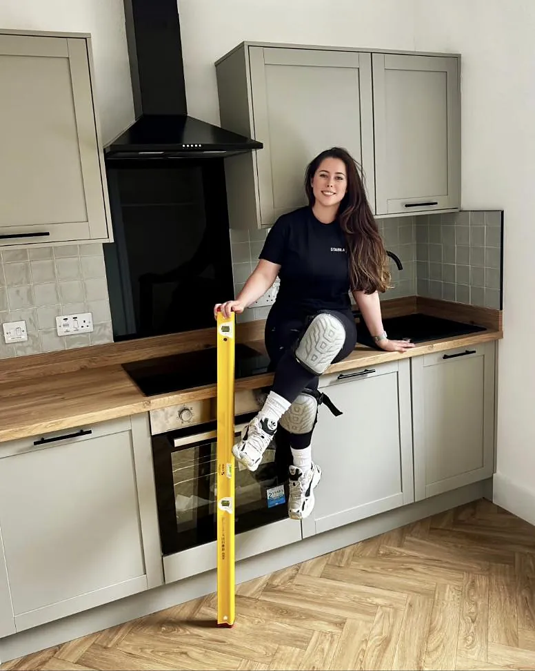 Emma sitting on the worktop of a grey shaker kitchen with a herringbone floor, holding a yellow spirit level